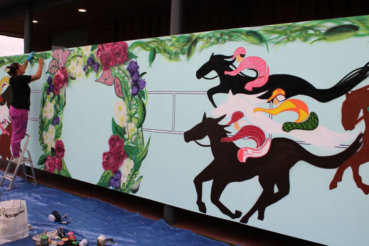 A woman on a ladder applying the finishing touches to a mural which features horse racing and a large decorative floral wreath for people to stand in front of.