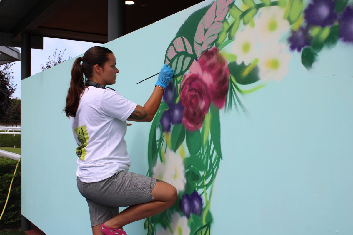 A side-on picture of a woman concentrating as she paints part of a large mural on a wall. The section of mural is a large wreath. The woman is standing on one leg.