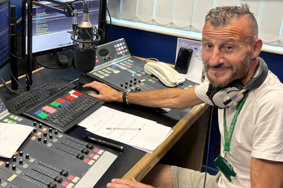 A radio presenter sat at the controls of a radio station, looking at the camera.