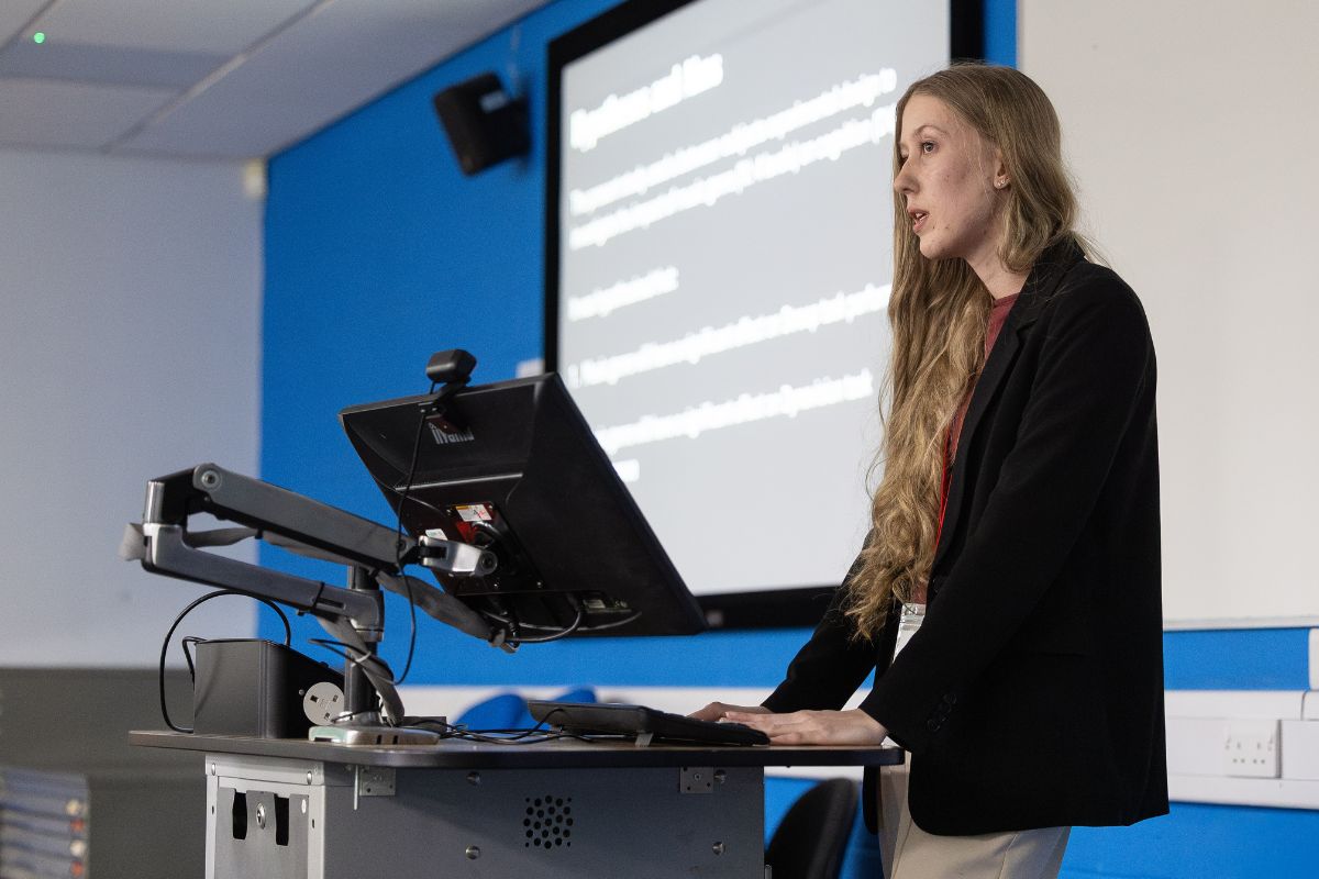 A woman with long hair giving a presentation standing at a desk by a computer monitor. In the background is a projected screen of text which is out of the camera