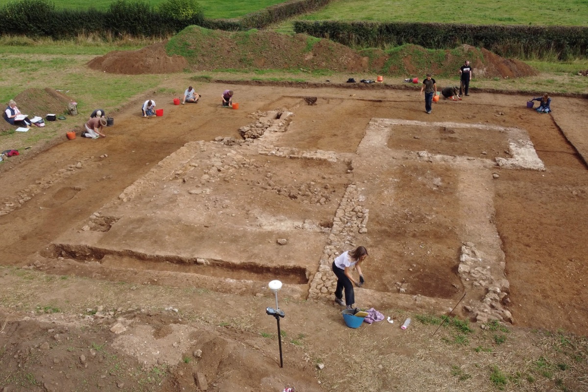 A photo taken from disrtance of people at work on an archaeological dig site. Stone work is exposed while a few small trenches have been dug. In the background is a mound of dug-up dirt and grass.