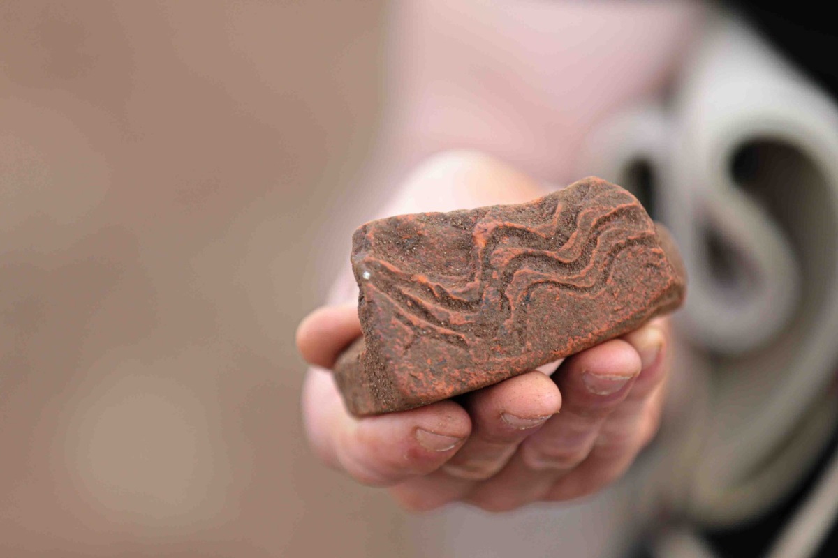 A close-up of an ancient piece of brickwork with wavy lines engraved on it. It is held in a hand. The rest of the photo is out of focus.