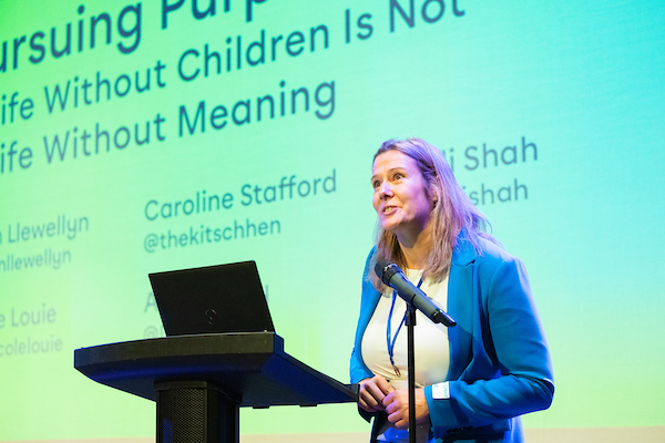 A woman at a lectern with a laptop, standing by a microphone, giving a lecture, in front of a big screen.