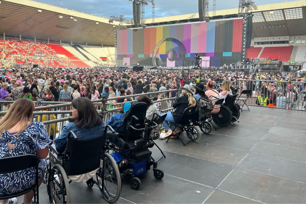 A large group of people gathered inside a stadium. In the front of the photo is a row of people attending the stadium event in wheelchairs and scooters.
