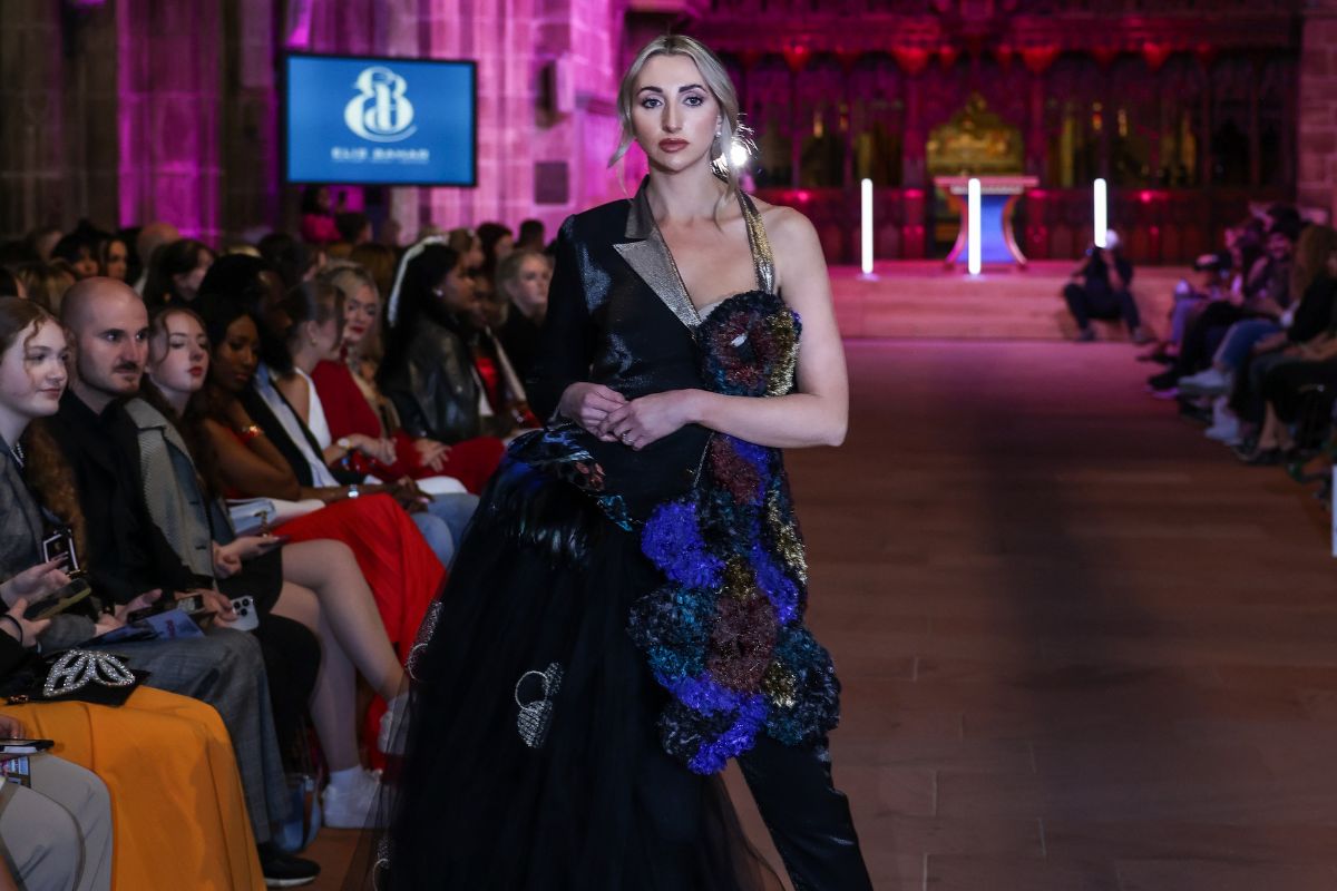 A fashion model in a designer outfit walking on a stone catwalk in front of a sitting crowd at Chester Cathedral.