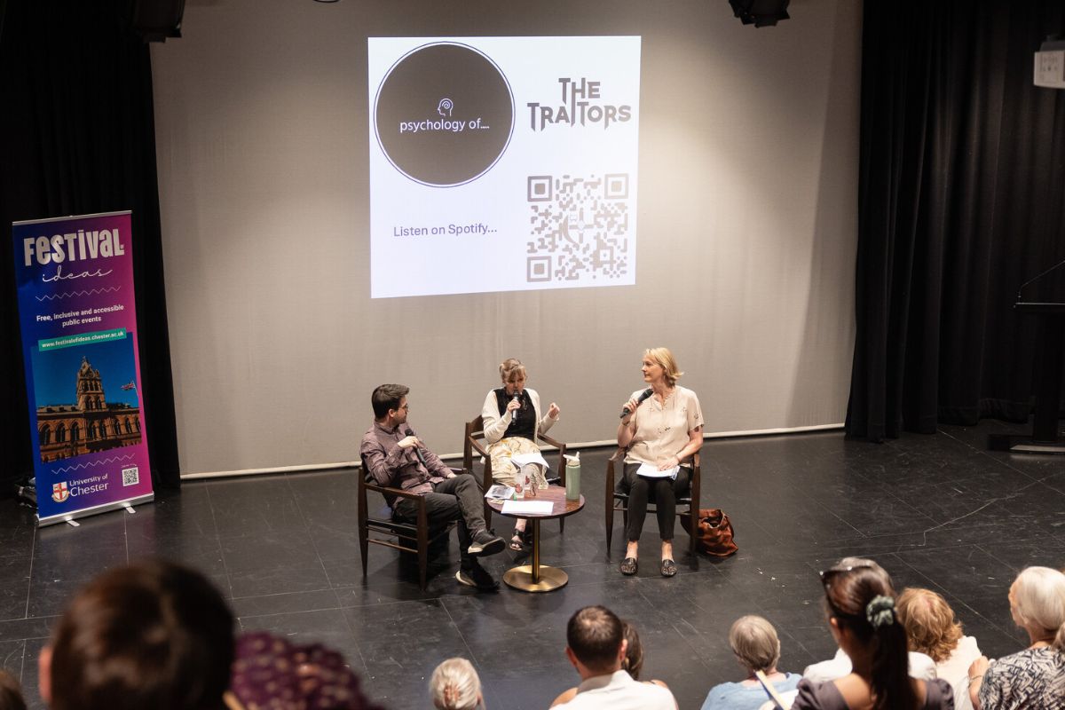 A man and two women sat down with microphones in a theatre space, with 'Psychology of... The Traitors' and 'Listen on Spotify' on the graphics in the background, along with a QR code. Some of the audience is visible in the photo.