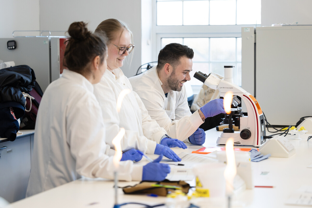 A group of people in a laboratory, smiling. Bunsen burners are lit. One of the people is looking into a microscope.