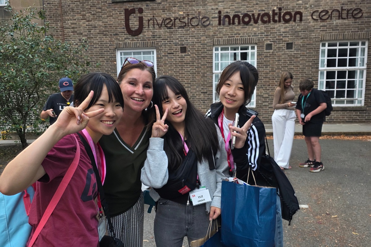 A group of students smiling and holding up two fingers in each hand while looking at the camera. In the background is the Riverside Innovation centre building, including its logo.