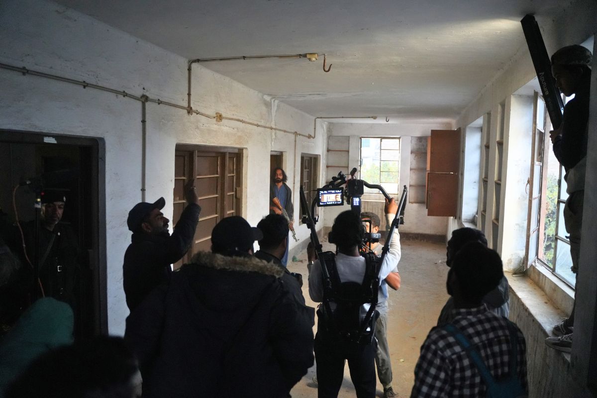 A film set with crew and cameras. The scene is a largely empty room. The film crew have their back to the camera.