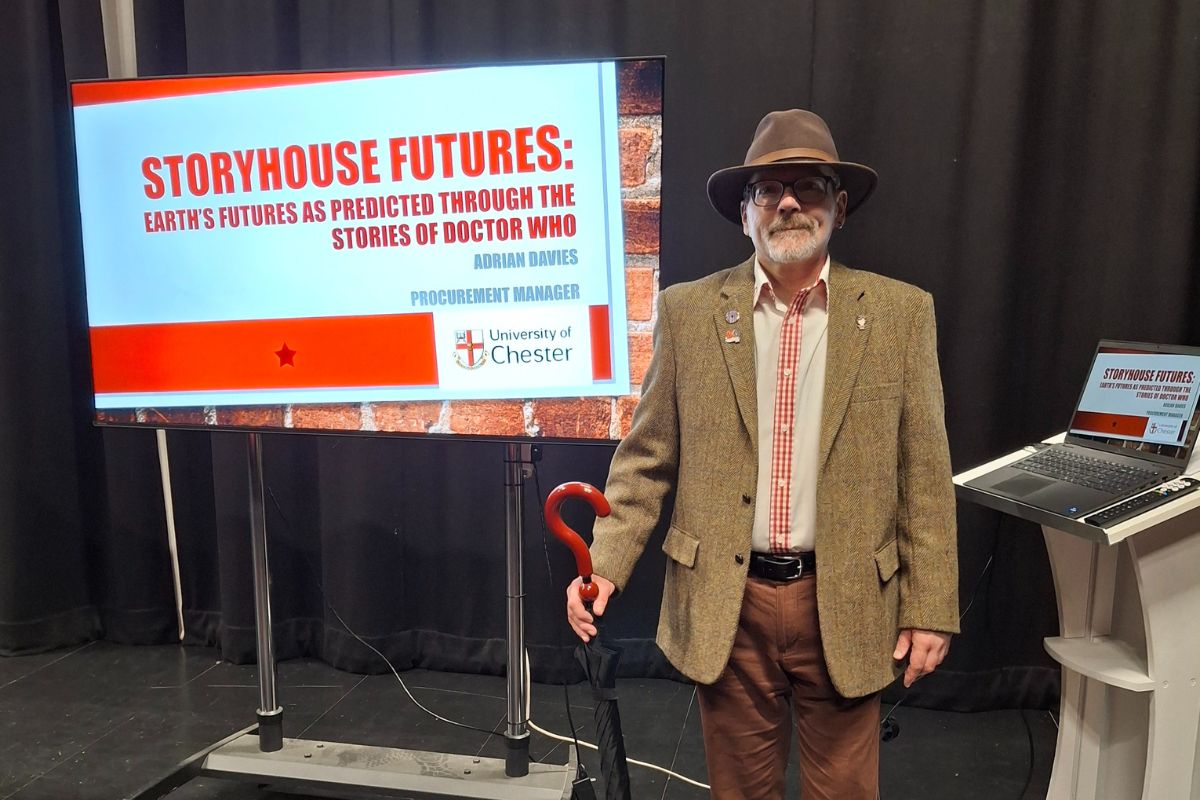 A man in a hat with a question-mark umbrella and blazer. The display in the background is titled 'Storyhouse Futures: Earth's futures as predicted through the stories of Doctor Who', by Adrian Davies, Procurement Manager, University of Chester.