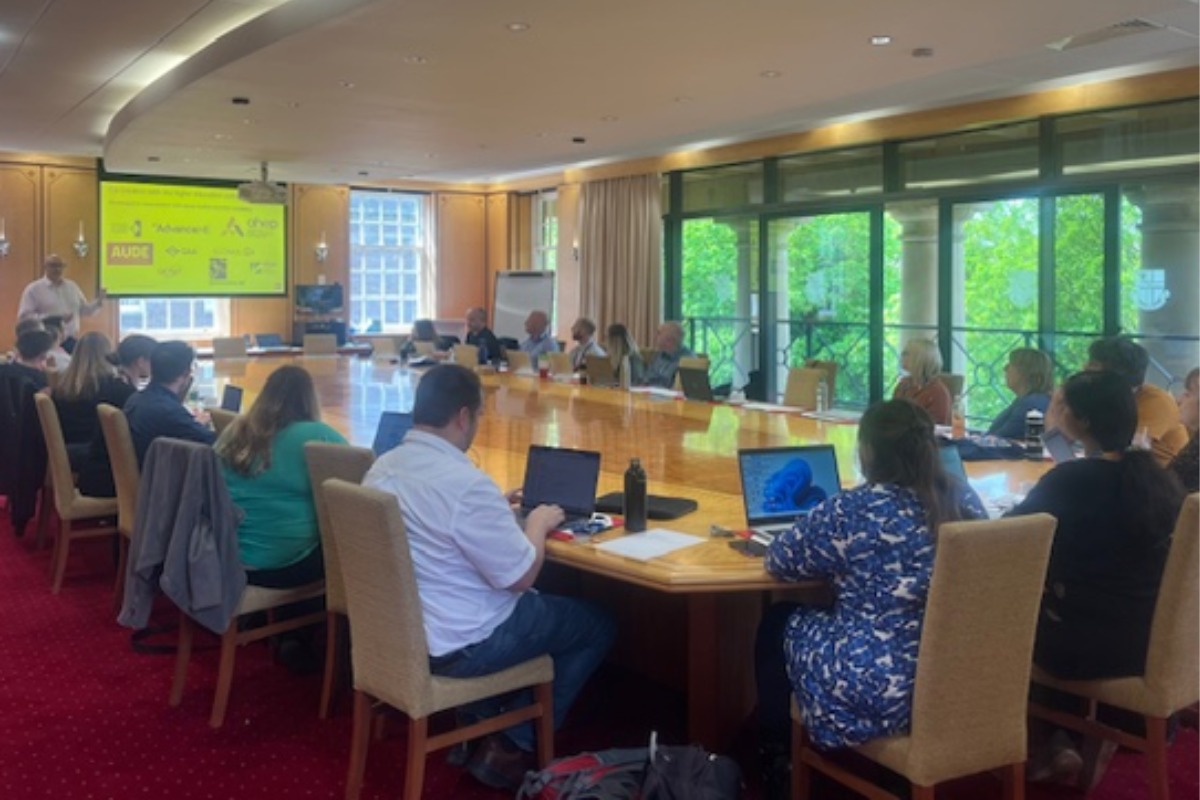 A group of people with laptops sitting around a table in a meeting with a slide shown as part of a presentation.