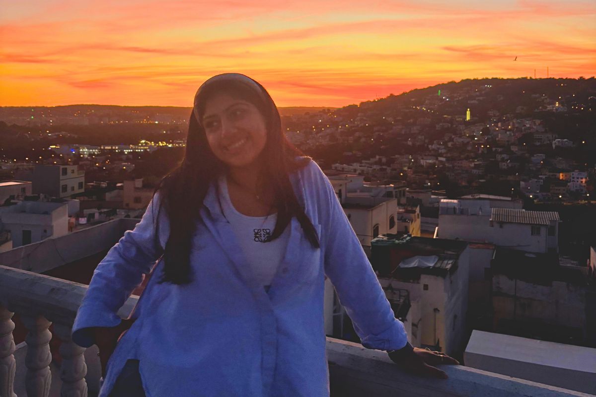 A woman at a balcony at sunset. The lighting is low. The woman is smiling and looking off camera. A cityscape is in the background.