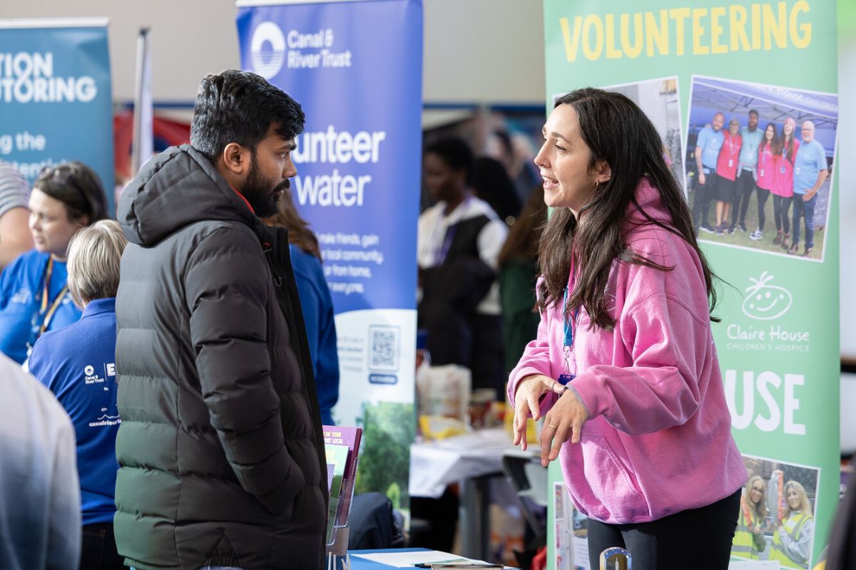 A student in a coat on the left listens to a stallholder woman on the right about one of the volunteering opportunities available.