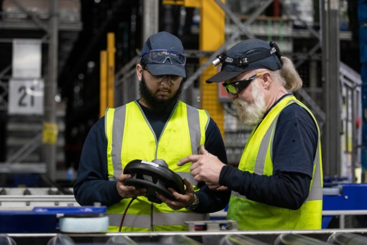 A student and a man in hi-vis jackets looking down on a piece of equipment in a factory setting.