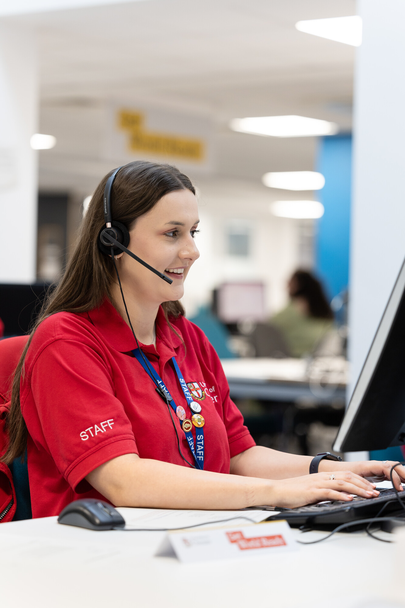 A woman sat in a chair with a headset on, smiling, typing on a computer keyboard