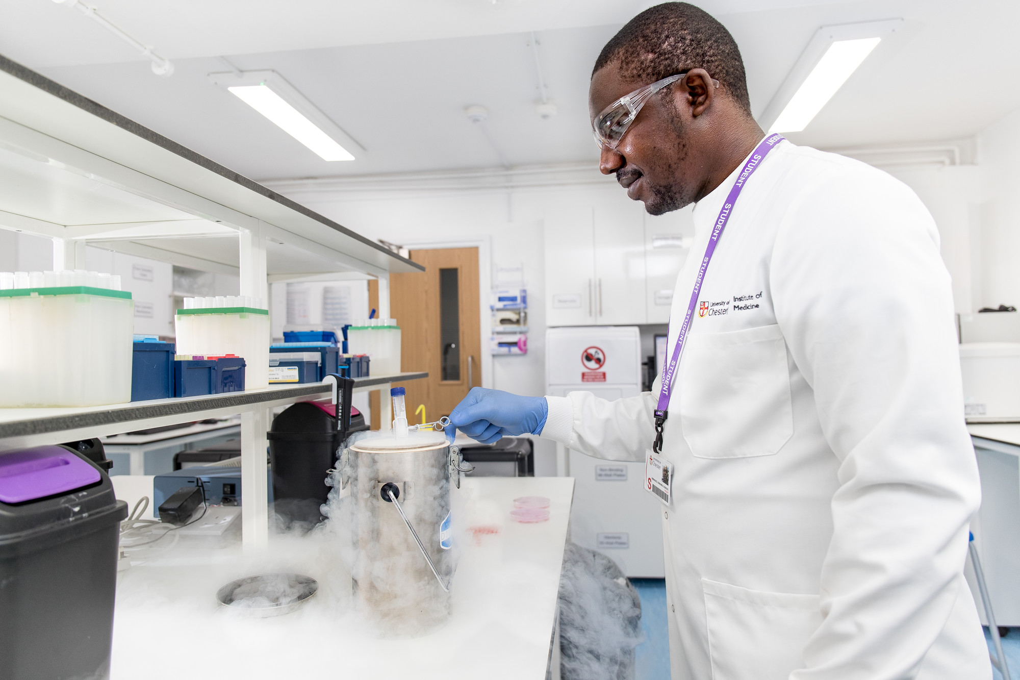A man with goggles and other lab safety equipment looks at a bubbling beaker.