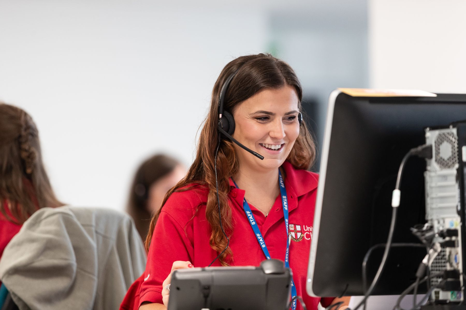 A woman in a red University of Chester top is wearing a headset and looking at a computer screen