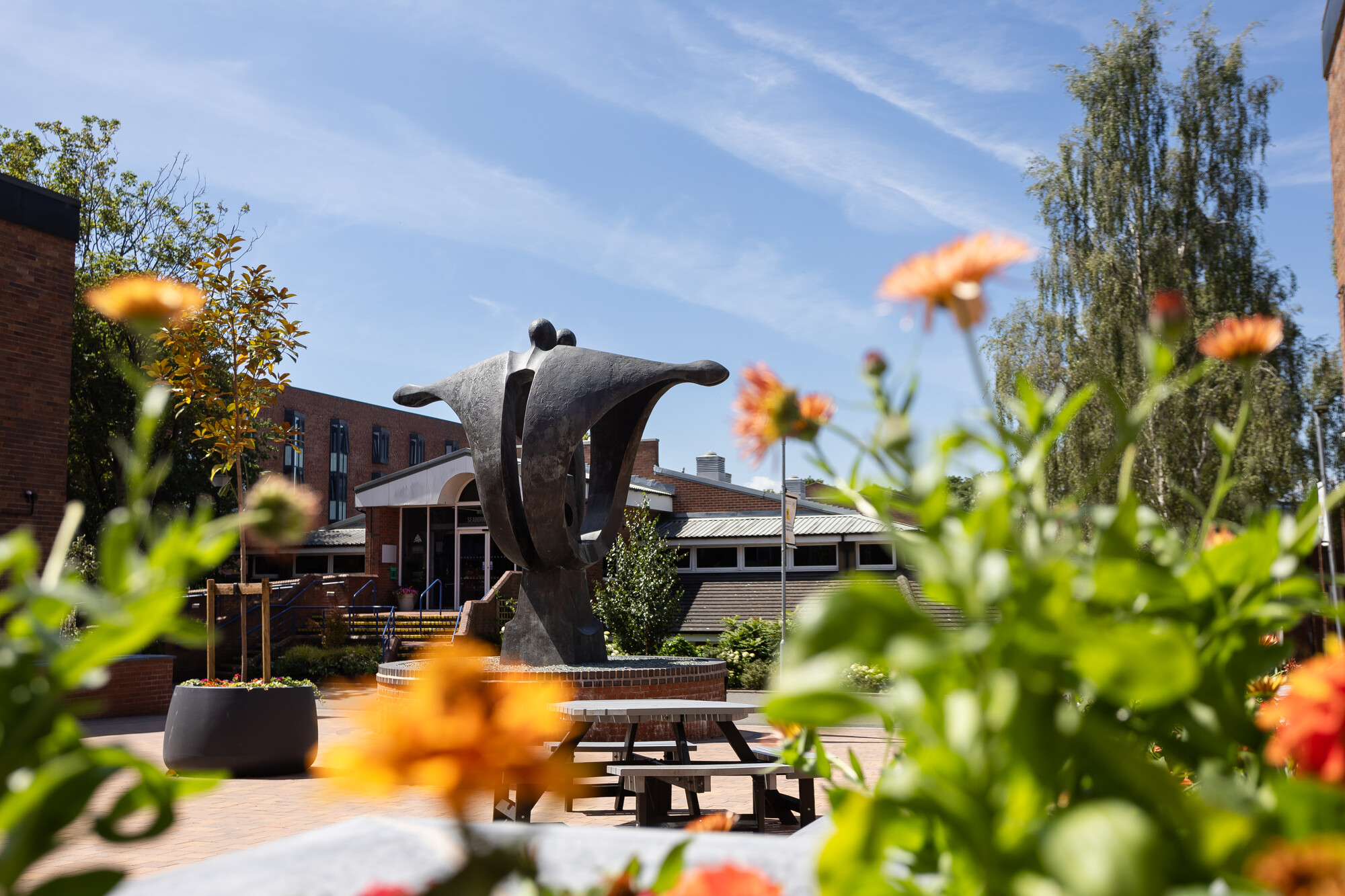 View of the University of Chester’s Exton Park campus, featuring the central sculpture framed by bright orange flowers, with the Seaborne Library and surrounding buildings in the background on a sunny day.
