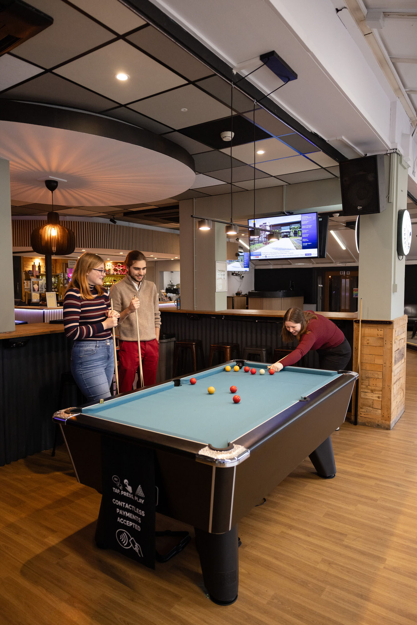 People playing a game of pool in a modern bar with a contactless‑payment pool table, wooden flooring, a large TV screen and contemporary interior décor.