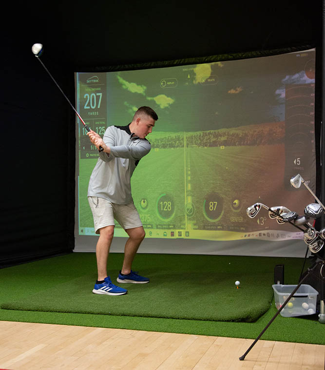 A man standing in front of virtual golf green screen practising his swing with golf club and ball.