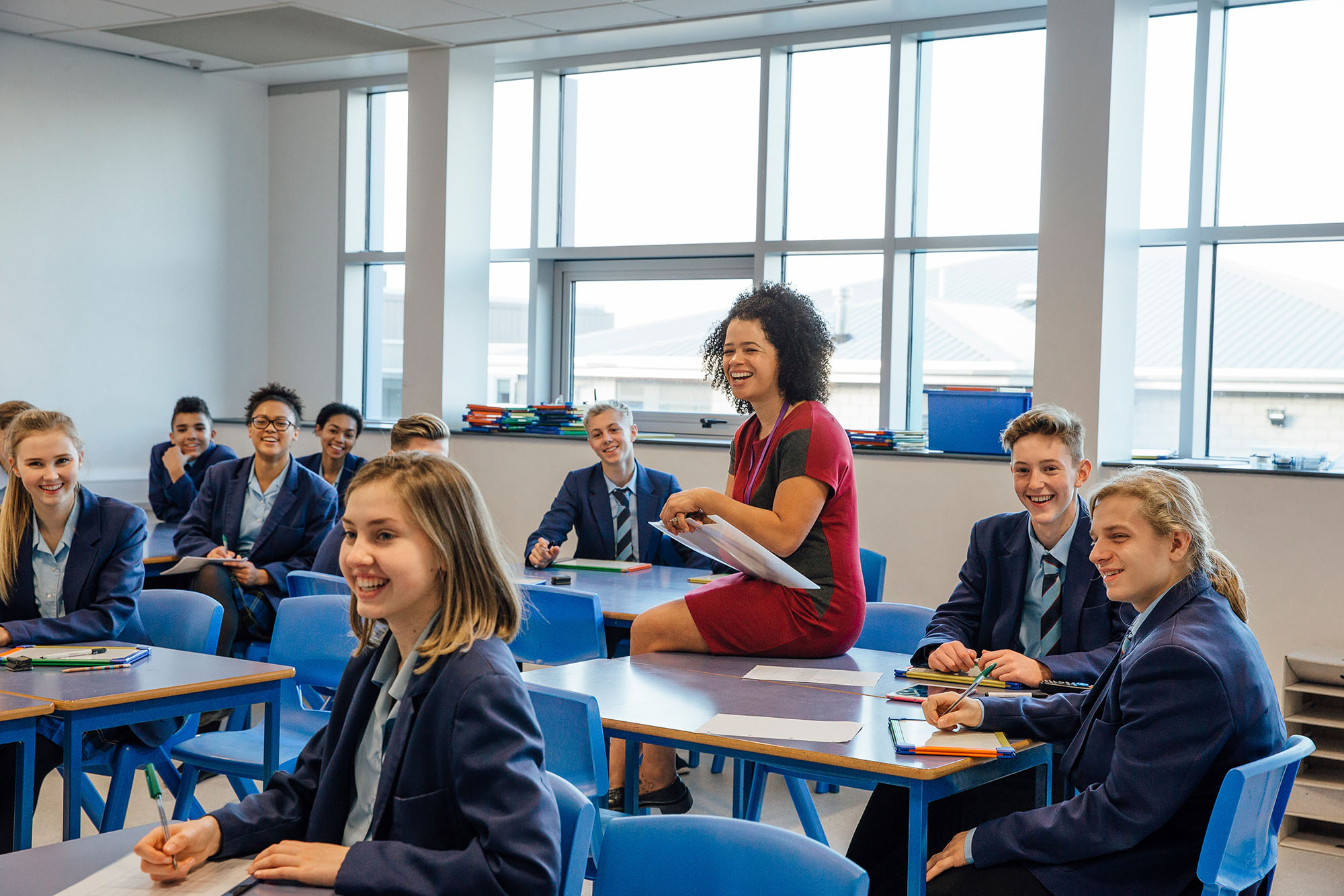 Happy secondary school students and a teaching assistant are laughing at their teacher who is out of the frame, during a school lesson.