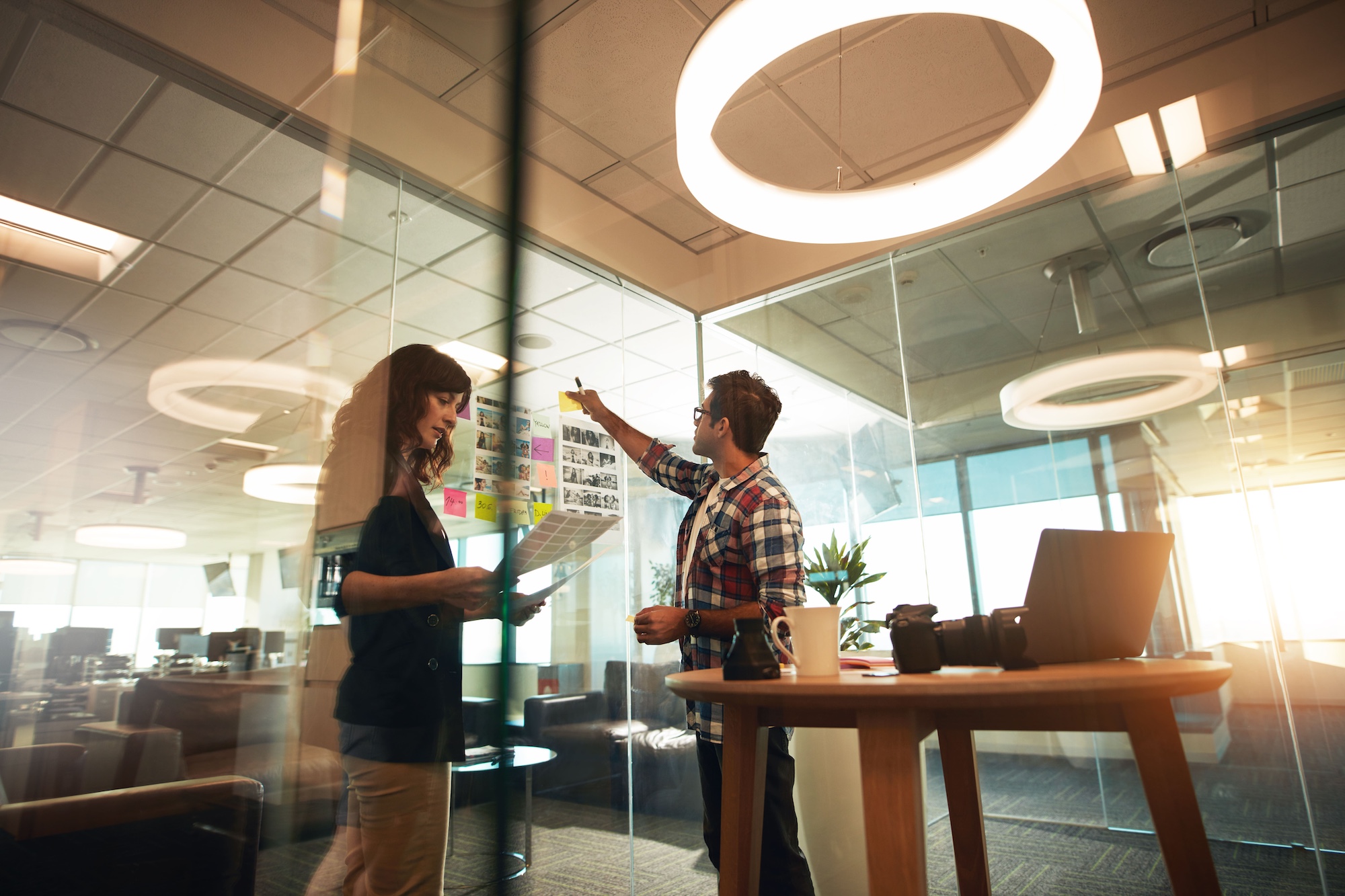 Two colleagues in an office with glass walls looking at sheets of paper and post it notes on a wall