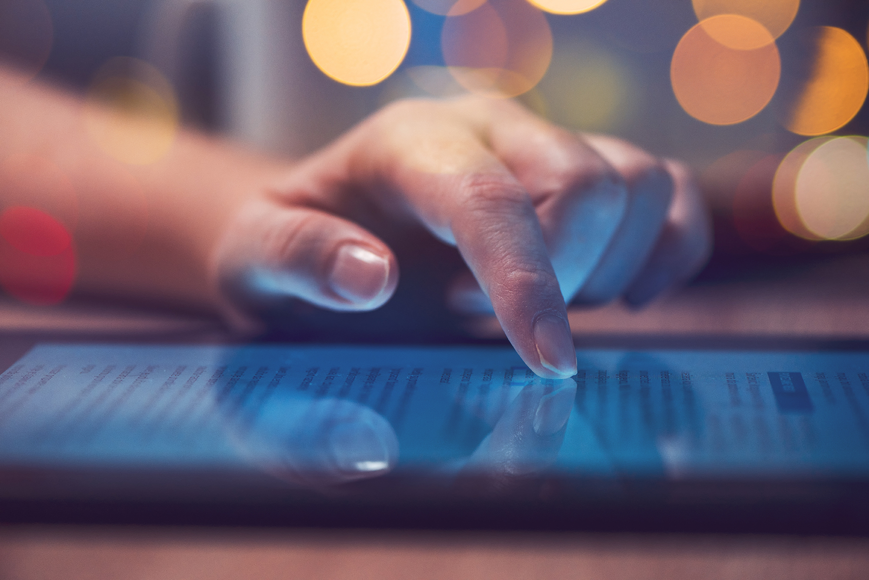 A close-up of a hand using a touchscreen tablet, with text visible on the screen and blurred colourful lights in the background.