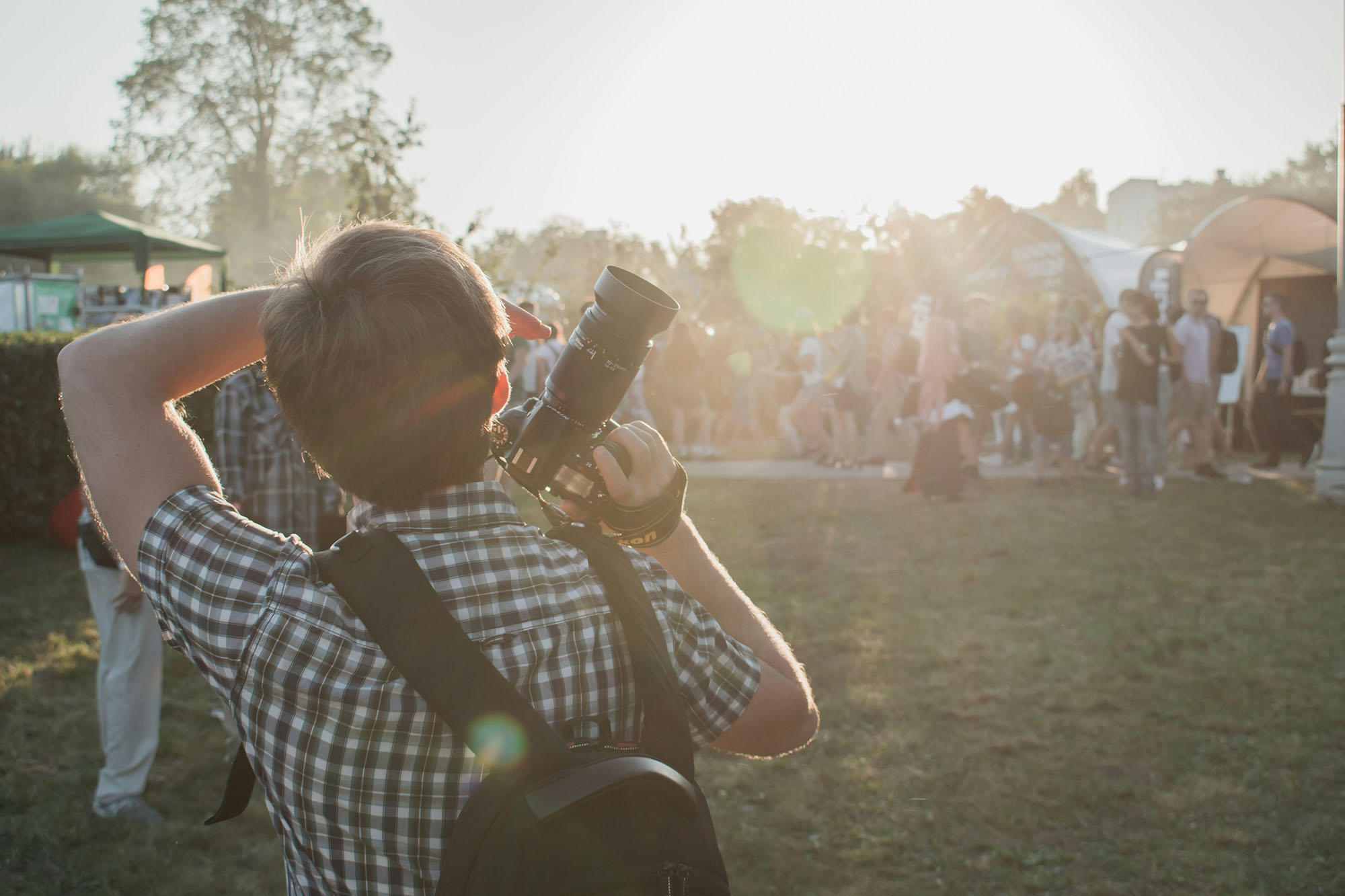 Man in grey and white plaid shirt with backpack and DSLR camera on hand
