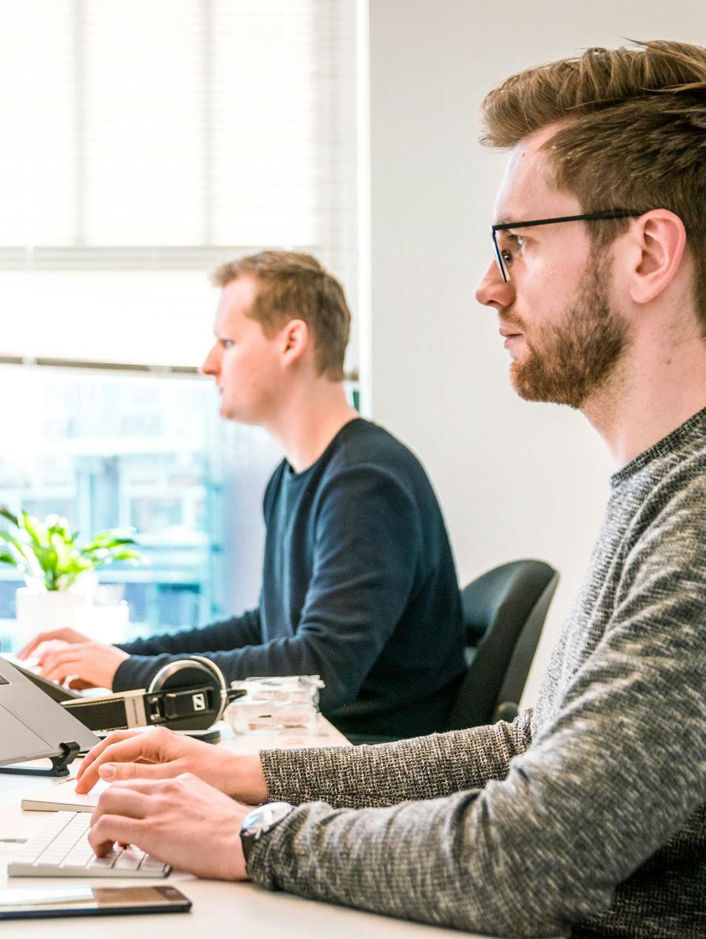 Two professionals working at modern computer workstations, analysing data and reviewing business insights on screen. The bright office setting conveys advanced research, strategic decision‑making, and evidence‑based practice—core themes of a Doctor of Business Administration (DBA) programme focused on leadership, innovation, and organisational performance.