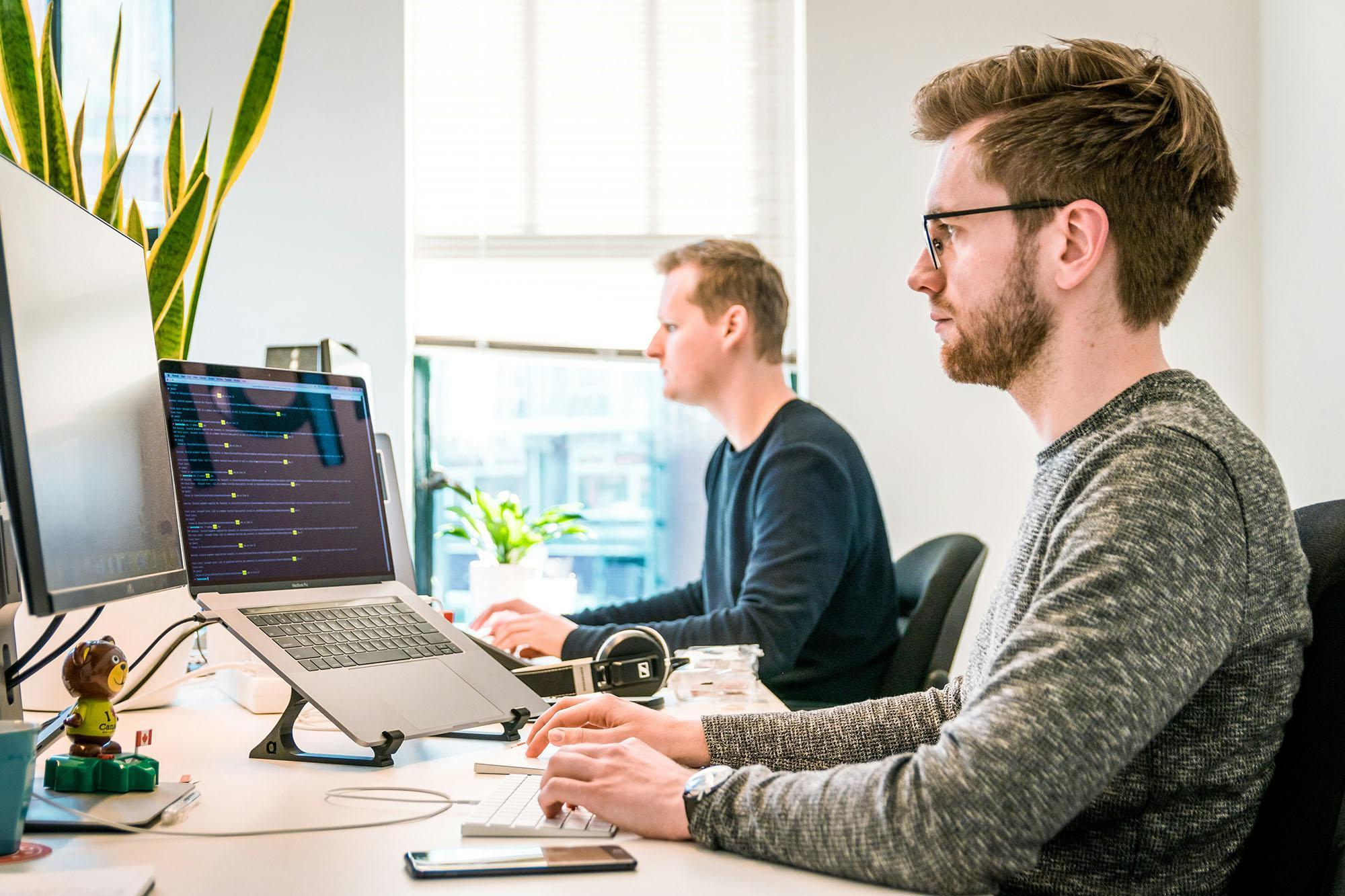 Two professionals working at modern computer workstations, analysing data and reviewing business insights on screen. The bright office setting conveys advanced research, strategic decision‑making, and evidence‑based practice—core themes of a Doctor of Business Administration (DBA) programme focused on leadership, innovation, and organisational performance.