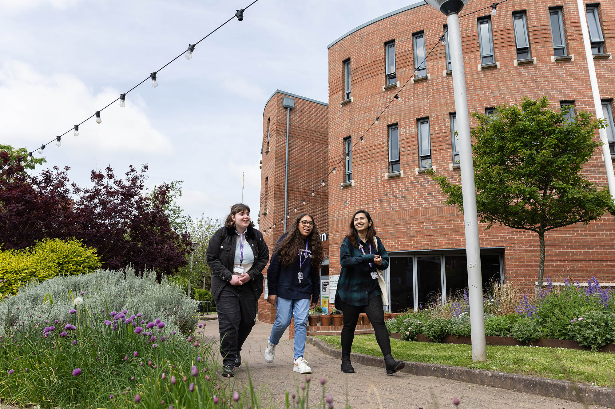 Three students walking together and smiling outside the Binks building at the University of Chester's Exton Park, surrounded by greenery and string lights overhead.