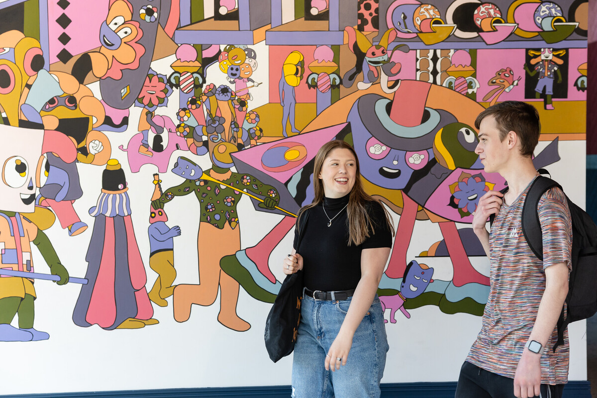 A male and female student, walking and talking in front of a colourful murial wall in Chester.
