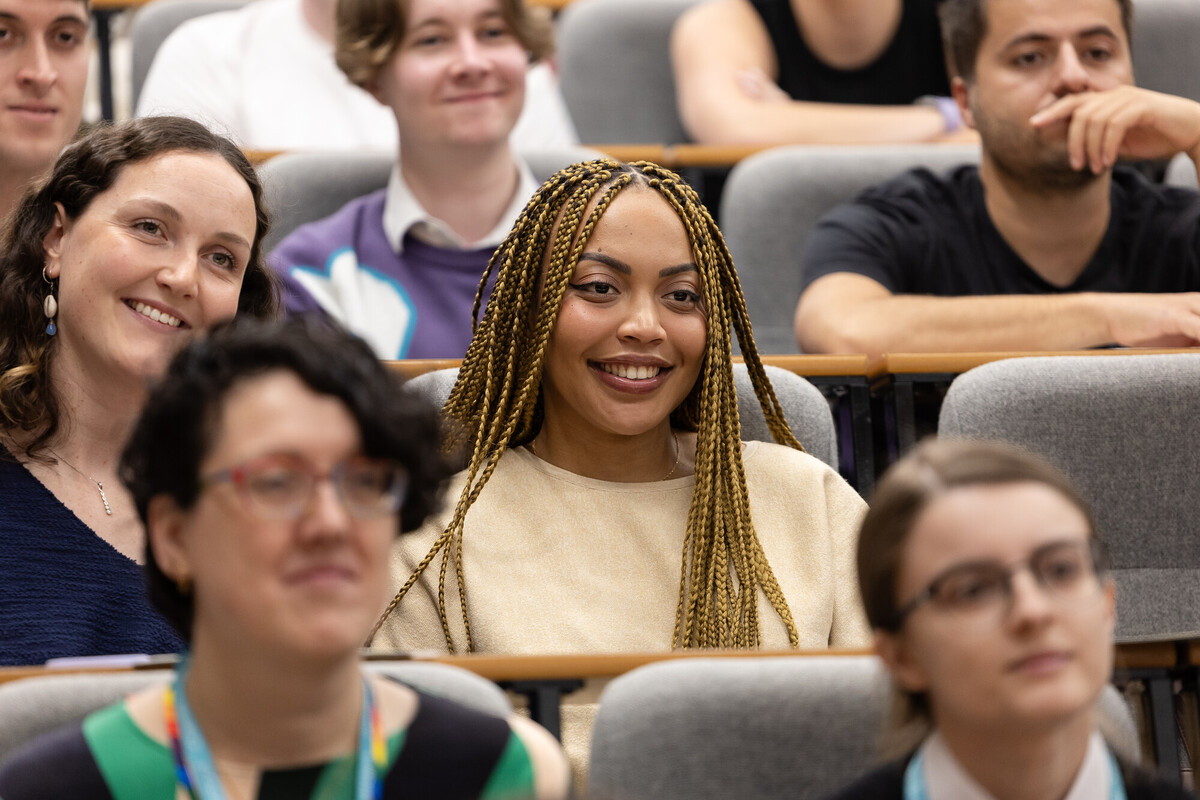 Students sitting in rows of a lecture theatre. Focus on one female, smiling and looking ahead
