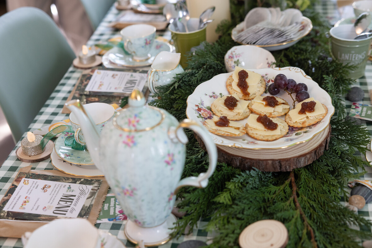 Decorated tea table with floral china, teapot, and a plate of crackers topped with cheese and chutney, surrounded by greenery and candles.