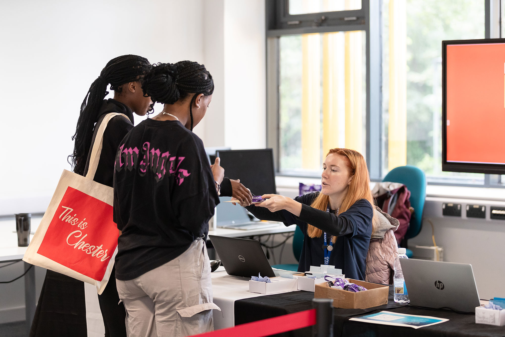 A student welcome or registration desk in a bright university building. Two students stand at the desk while a staff member reaches across to hand over a lanyard or information item. A tote bag with the words “This is Chester” hangs from one student’s shoulder. Laptops, event materials and a display screen are visible around the check-in area.