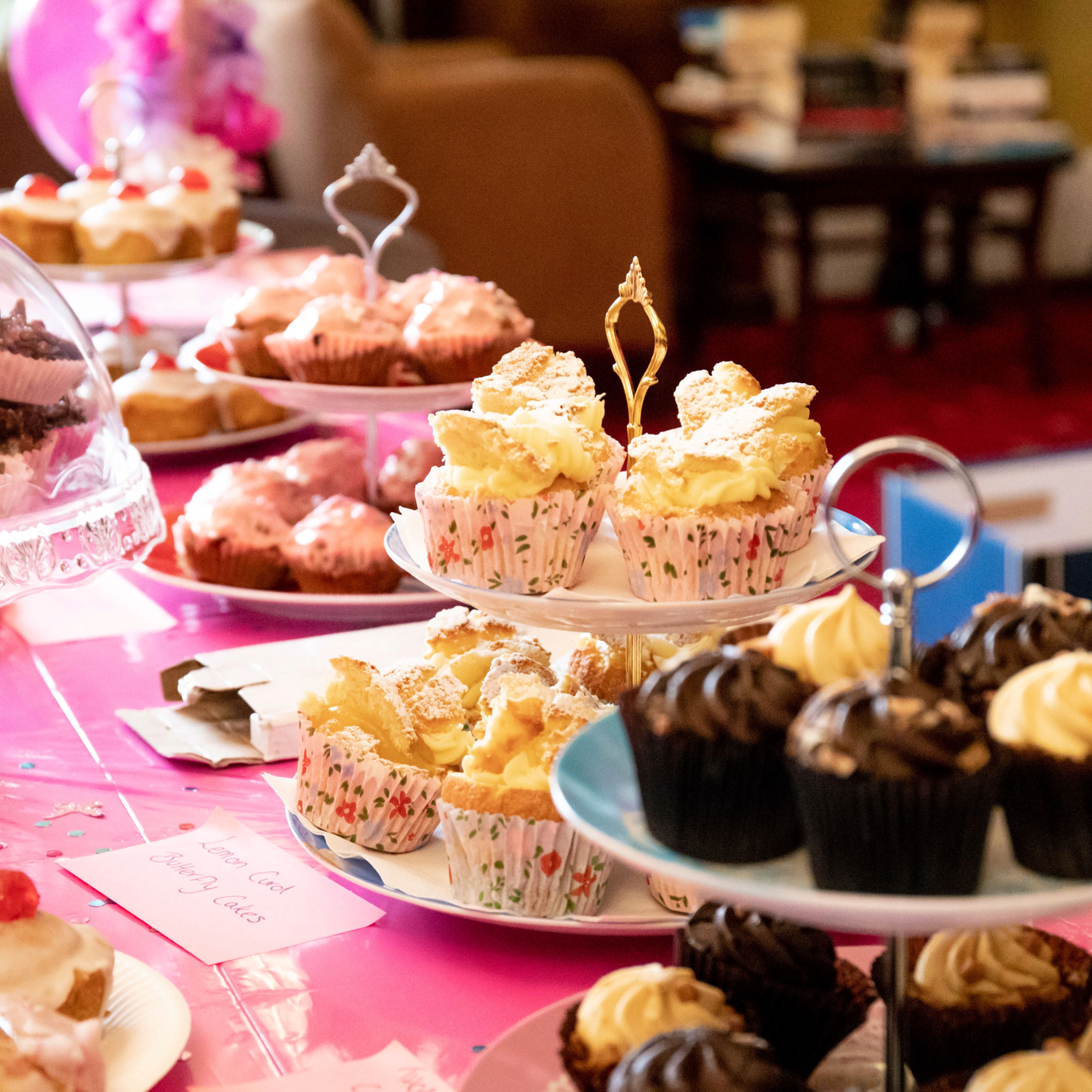 Selection of homemade cakes and cupcakes displayed on tiered stands on a pink-decorated table at a bake sale.